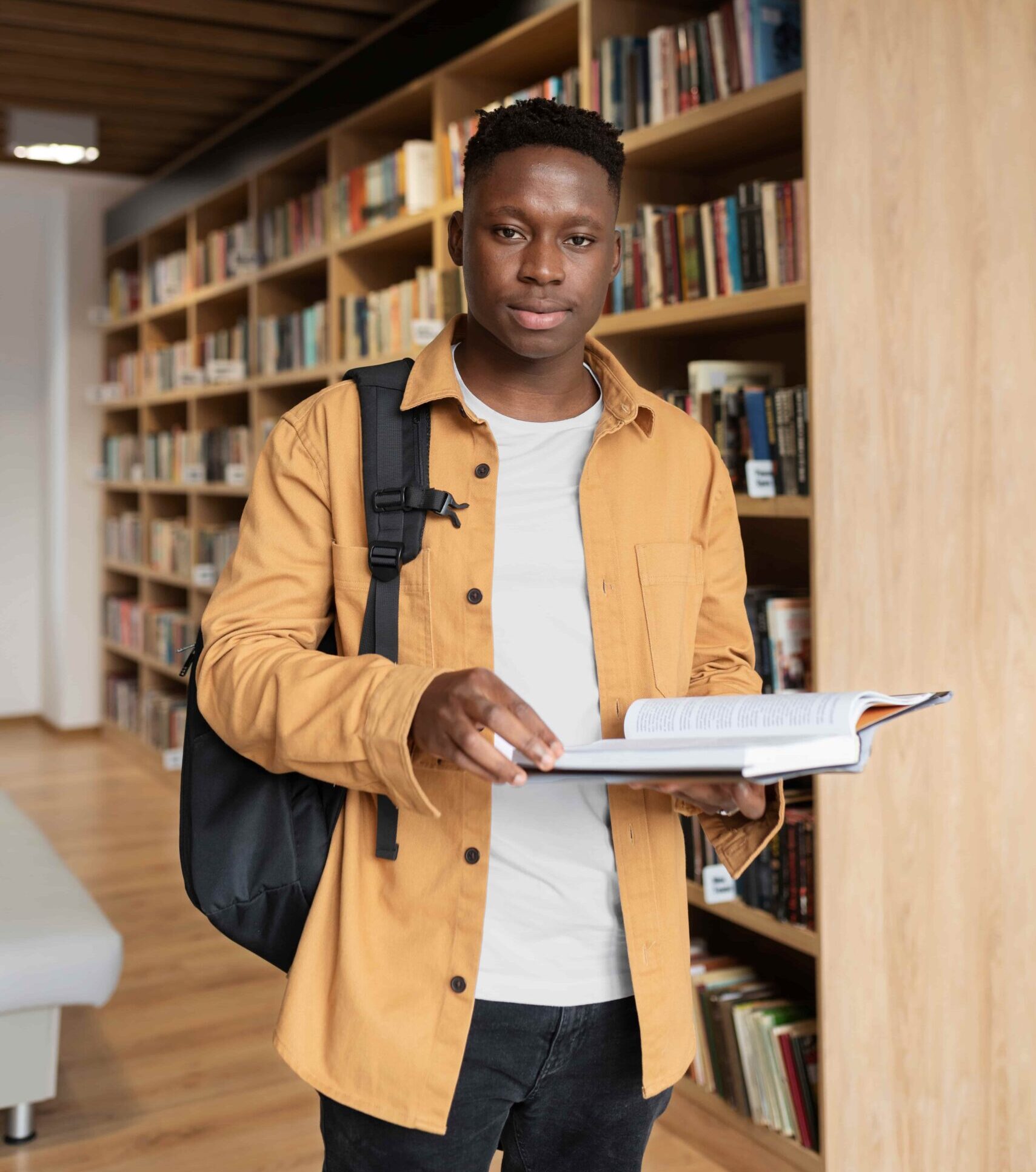 Focused student wearing a mustard jacket and backpack, reading an open book in a bright modern library with wooden shelves and exposed ceiling beams.